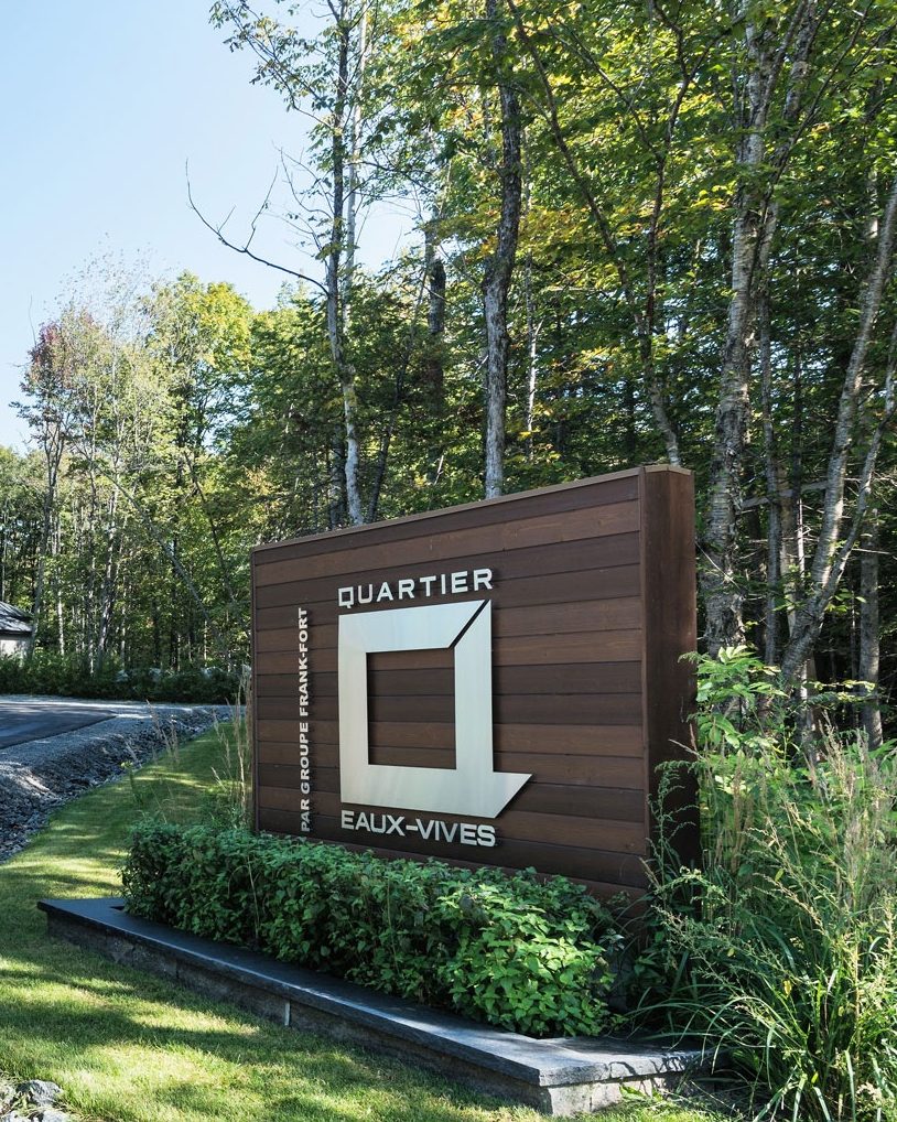 L'enseigne de bois du Quartier Eaux-Vives, avec son logo, devant la forêt