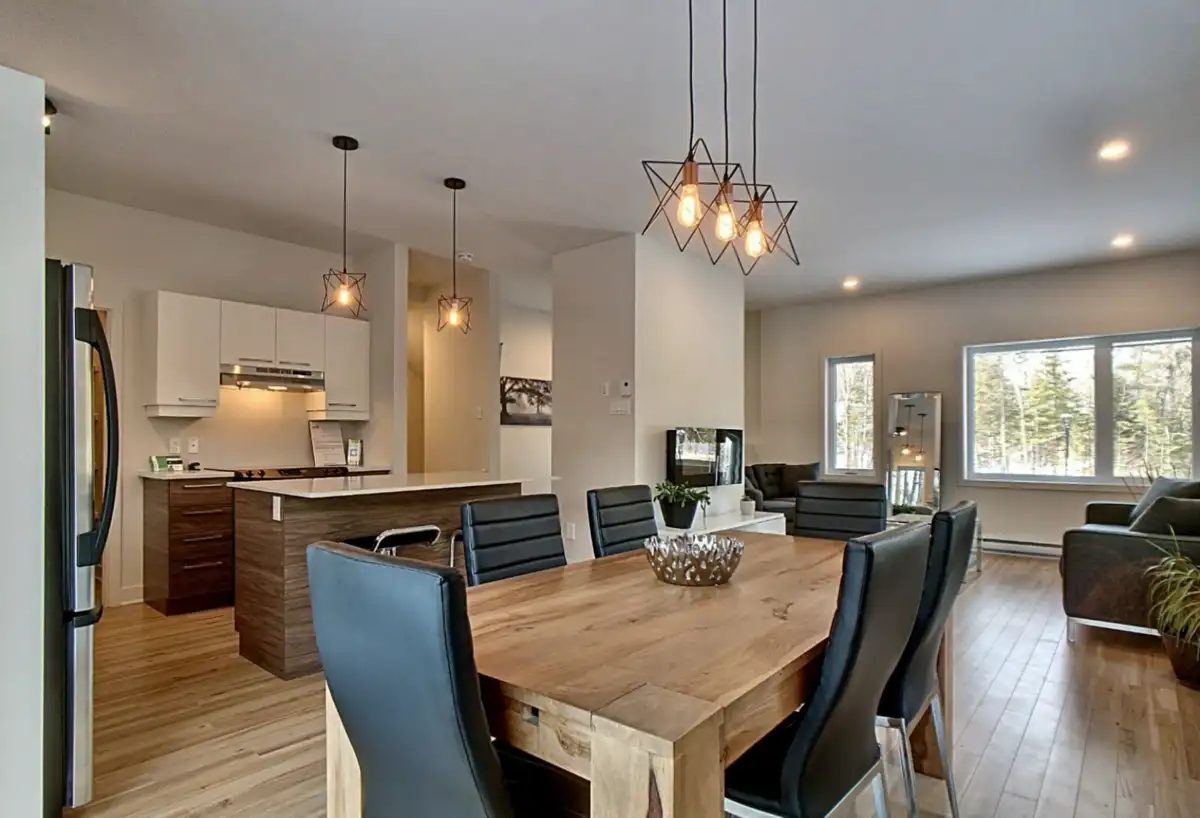 Intérieur de la maison modèle du Quartier Eaux-Vives : cuisine ouverte, grande table de bois, luminaires suspendus, le salon et les fenêtres sur le boisé au fond