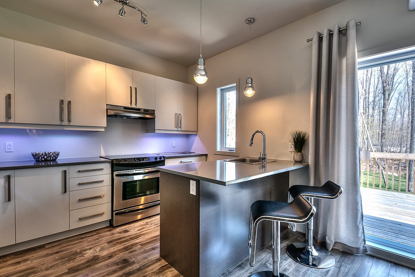 Cuisine blanche au Quartier Eaux-Vives : dosseret éclairé, cuisinière en acier inoxydable, porte-fenêtre sur la galerie